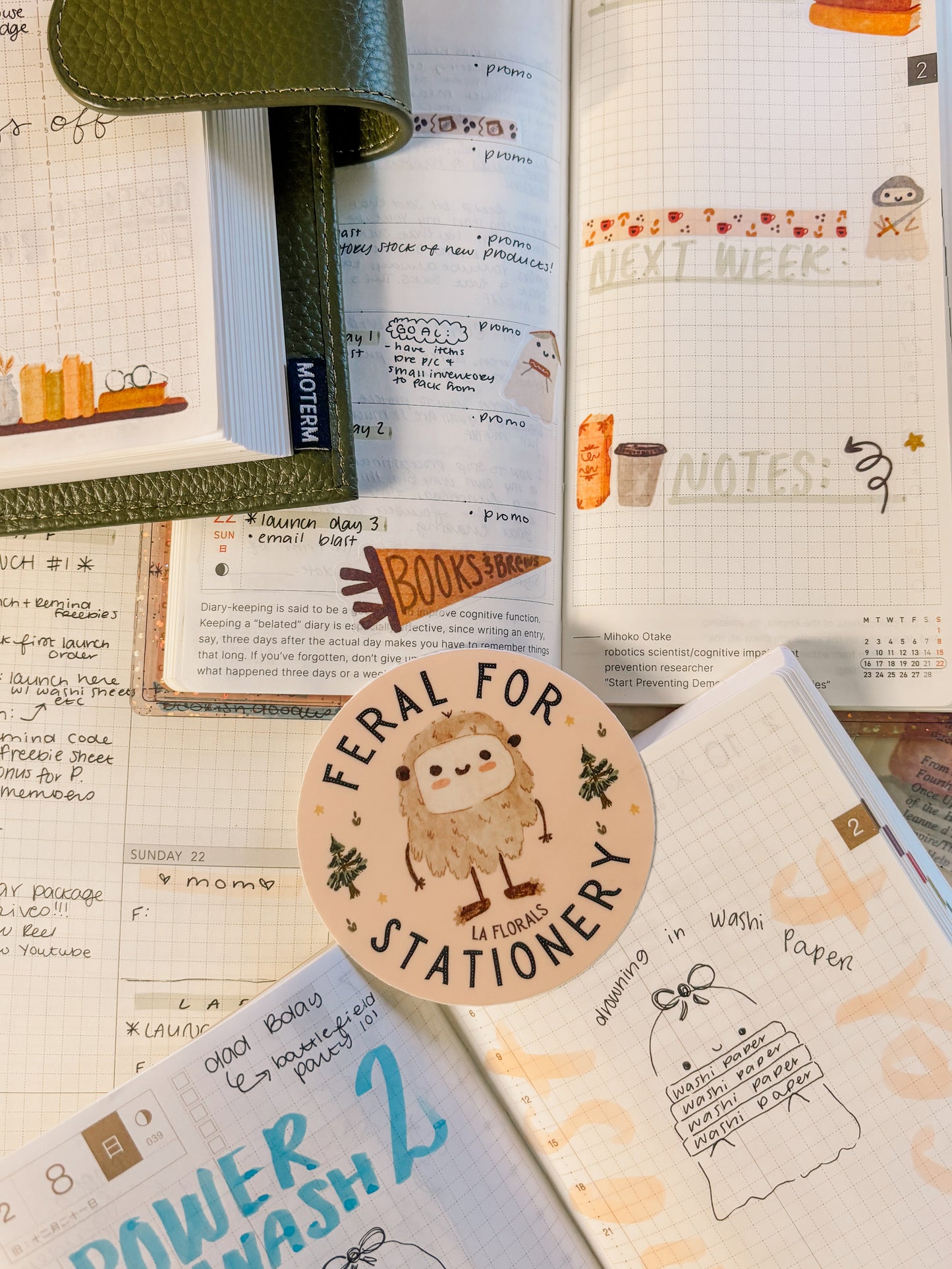 A vinyl sticker featuring a cartoon Bigfoot rests on a bed of open planners and notebooks. The sticker is a pale pink circle, and it also features the words, "Feral for Stationery - LA Florals."